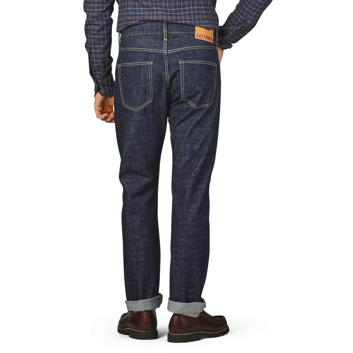 A person is standing facing away, wearing a blue checkered shirt tucked into C.O.F Studio's Blue 14oz Kuroki Selvedge Cotton M8 Jeans with a leather patch on the back pocket, and brown shoes.