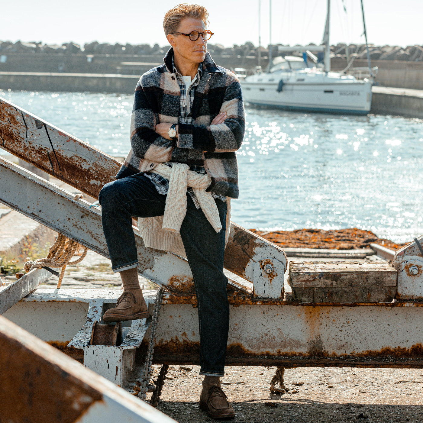 A man wearing C.O.F Studio's Blue 13oz Kuroki Selvedge Cotton M5 Jeans is sitting on a railing next to a boat.