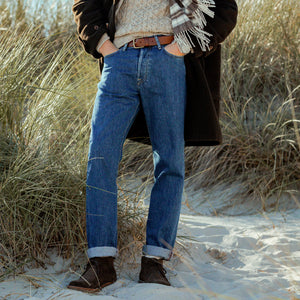 A man wearing a tailored fit coat stands confidently on the sand, his attire perfectly complemented by a pair of C.O.F Studio Blue Organic Candiani Cotton M6 6x Wash Jeans.