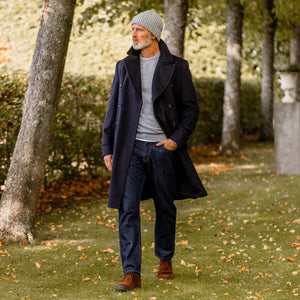 A man in a navy peacoat walking through a park wearing C.O.F Studio's Blue Rinsed Organic Candiani Cotton M7 Jeans.