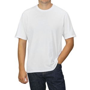 A person dressed in a C.O.F Studio White Organic Cotton Crewneck T-shirt and dark jeans, standing against a neutral grey background with one hand in their pocket.