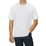 A person dressed in a C.O.F Studio White Organic Cotton Crewneck T-shirt and dark jeans, standing against a neutral grey background with one hand in their pocket.