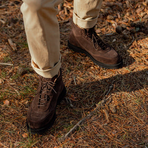 A person wearing CQP Dark Brown Suede Leather Sabulo Boots and beige pants stands on a forest floor covered with pine needles.