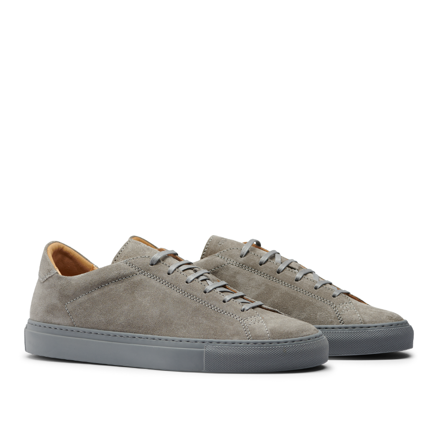 A pair of Granit Grey Suede Leather Racquet Sr Sneakers by CQP with white laces and a dark gray sole, displayed against a striped gray background.