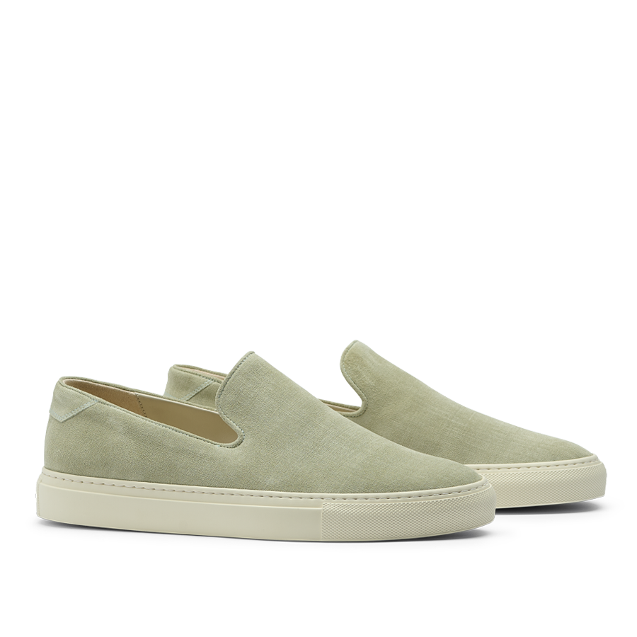 The CQP Sage Green Textured Suede Jetty Wholecut Slip-On sneakers feature a sleek slip-on design with crisp white soles, handmade in Portugal. These shoes showcase effortless style and quality craftsmanship when viewed from the side.