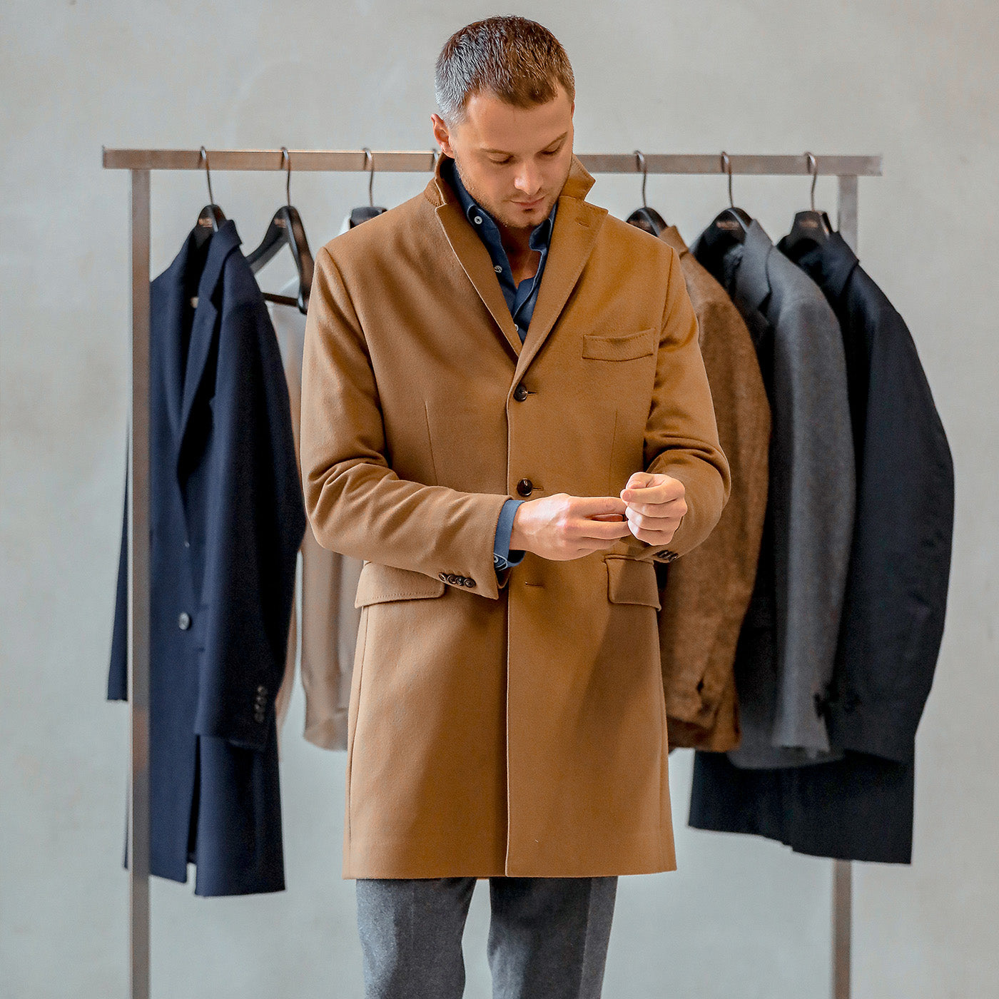 A man adjusts his cuff while wearing the Camel Beige Loro Piana Wool Tailored Coat by Baltzar Sartorial, in front of a clothing rack with several coats hanging, including exclusive pieces from Loro Piana.