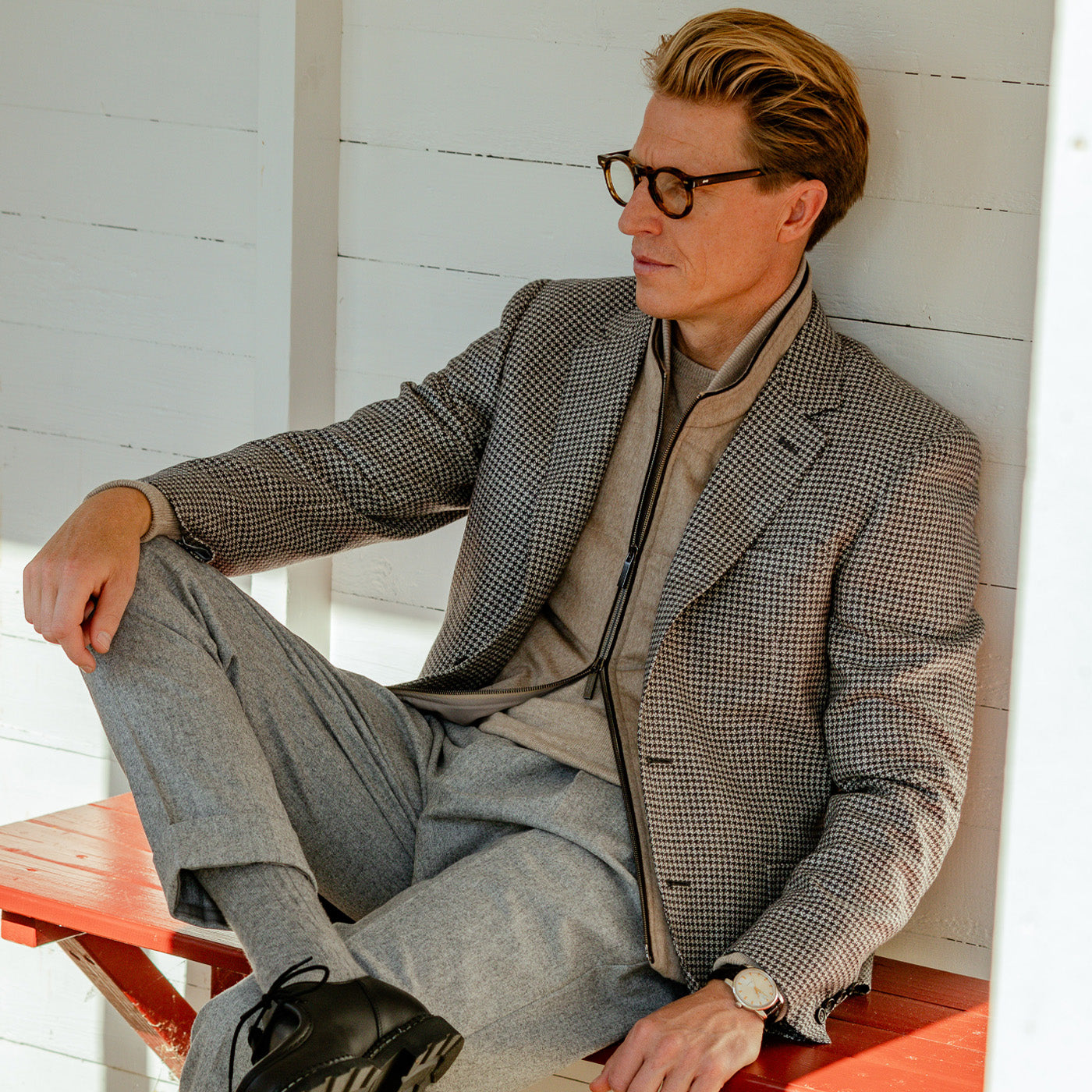 A man sitting on a bench wearing glasses and a Canali Grey Houndstooth Wool Drop 6 Blazer.