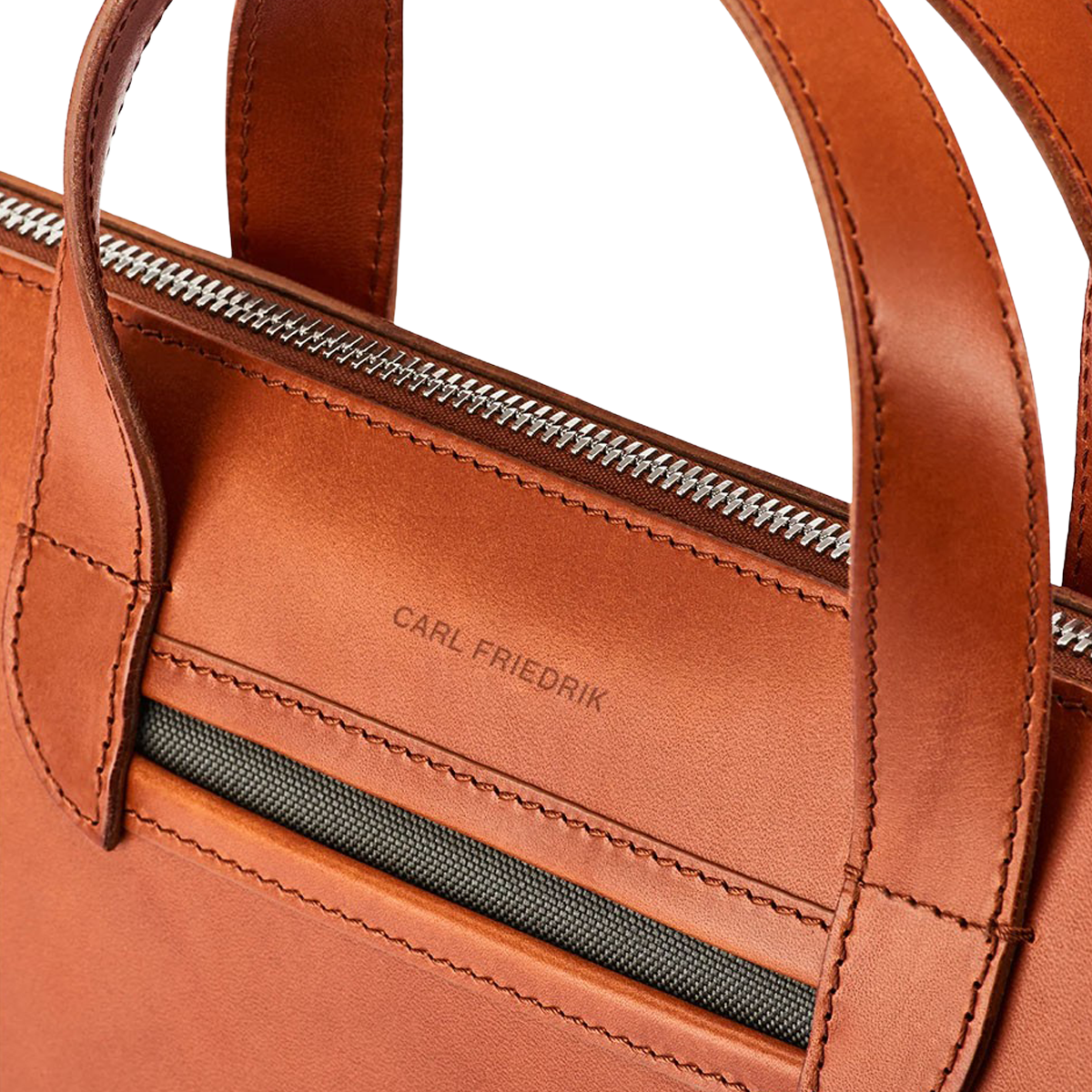 Close-up of a Cognac Vachetta Leather Vallance Slim Briefcase made from Italian vachetta leather with visible stitching, dual handles, a zipper closure, and the brand name "Carl Friedrik" embossed on the front pocket.