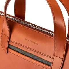 Close-up of a Cognac Vachetta Leather Vallance Slim Briefcase made from Italian vachetta leather with visible stitching, dual handles, a zipper closure, and the brand name "Carl Friedrik" embossed on the front pocket.