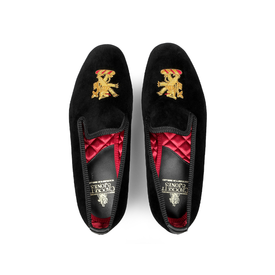 A pair of Black Velvet Lion Rampant Albert Slippers by Crockett & Jones, featuring hand-embroidered gold crests, a red quilted interior lining, and durable leather soles.