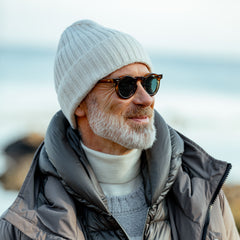 A man with a gray beard is smiling, wearing sunglasses, the Nuvola Ecru Ribbed Cashmere Beanie by Daniele Fiesoli, and a gray puffer jacket made from upcycled cashmere. The background is a blurred outdoor setting.