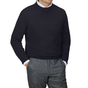 A person stands against a gray backdrop, stylishly wearing the Navy Blue Cashmere Crewneck Sweater by Daniele Fiesoli over a white collared shirt, paired with gray pants.