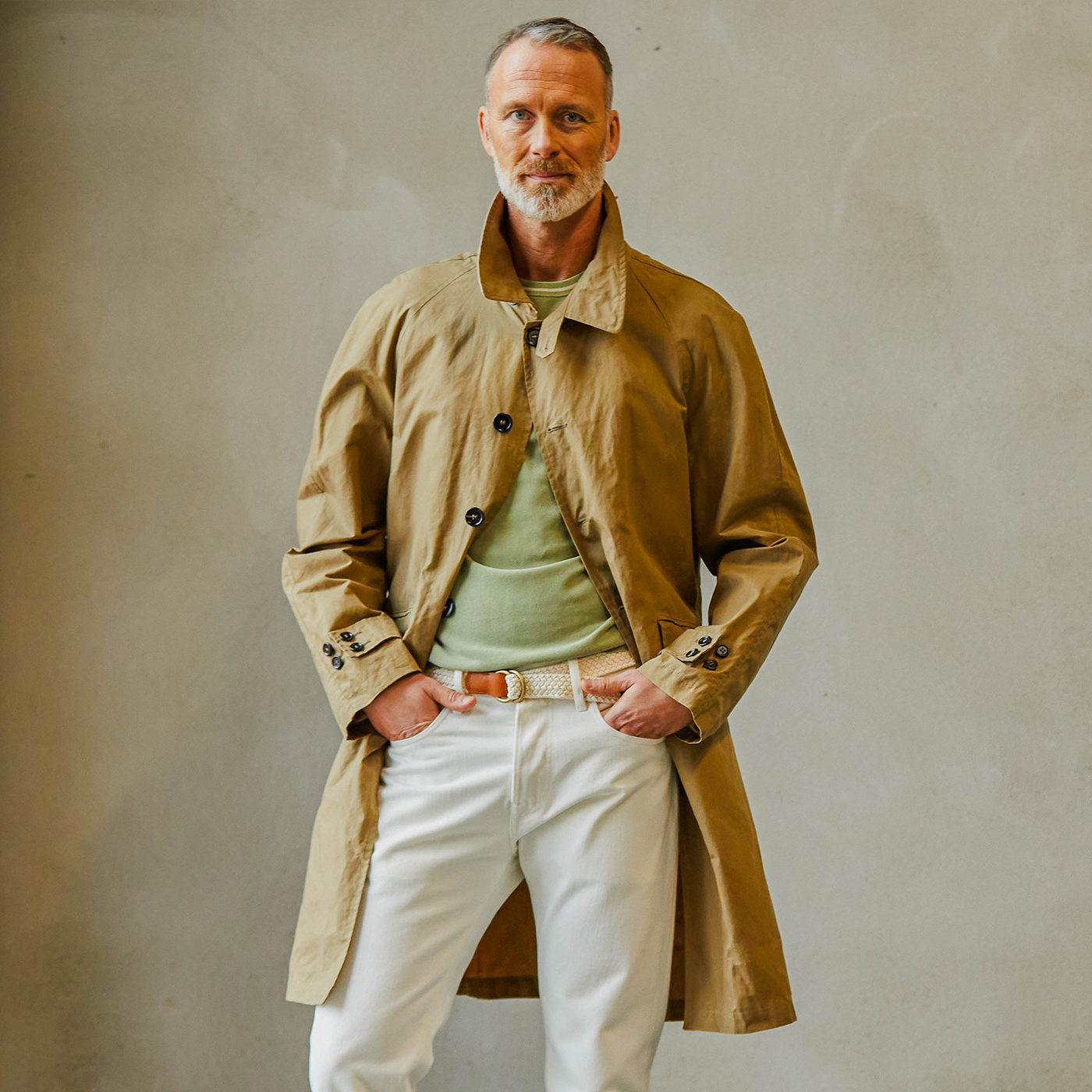 A man wearing a dark beige Waxed Cotton Kamikaze trenchcoat by L'Impermeabile over a light green sweater and white pants, standing with his hands on his hips and giving a confident look.
