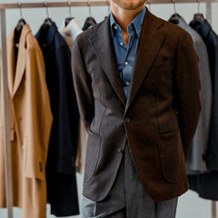 A man wearing a dark brown wool tweed blazer by Baltzar Sartorial and a blue shirt stands in front of a rack of various colored jackets, some possibly crafted from the renowned Abraham Moon collection.