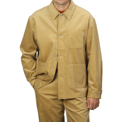 A person wearing the Gold Mustard Cotton Corduroy Work Jacket from De Bonne Facture with a collar and large front pockets, matching tan pants, and an orange shirt underneath, against a plain gray background.