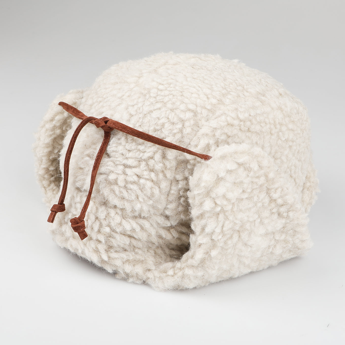 The Natural Wool Fleece Chapka Hat by De Bonne Facture showcases fluffy, white natural wool fleece and is accented with elegant brown calf leather strings, all set against a plain white background.