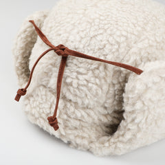 Close-up of a fluffy, cream-colored Natural Wool Fleece Chapka Hat by De Bonne Facture, featuring ear flaps tied together with a sleek calf leather string.