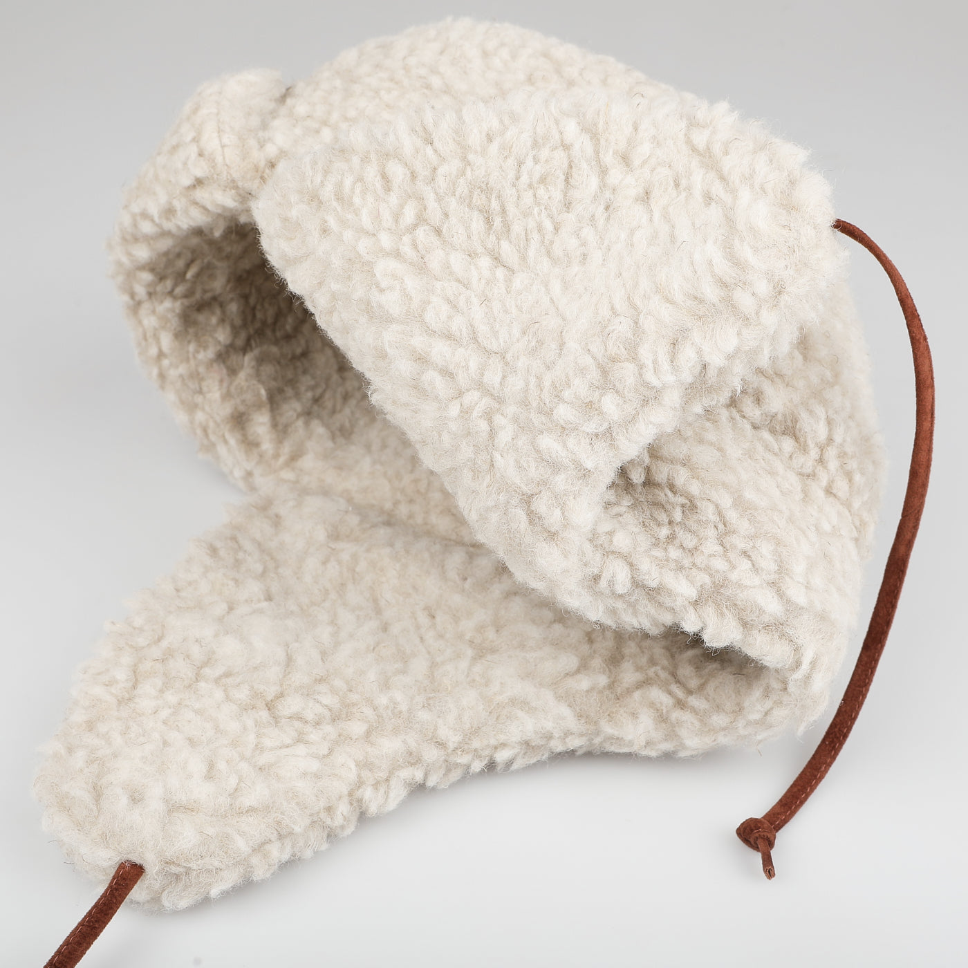 The Natural Wool Fleece Chapka Hat by De Bonne Facture features a cream-colored, fluffy design with ear flaps made from natural wool fleece and brown calf leather strings, set against a light gray background.