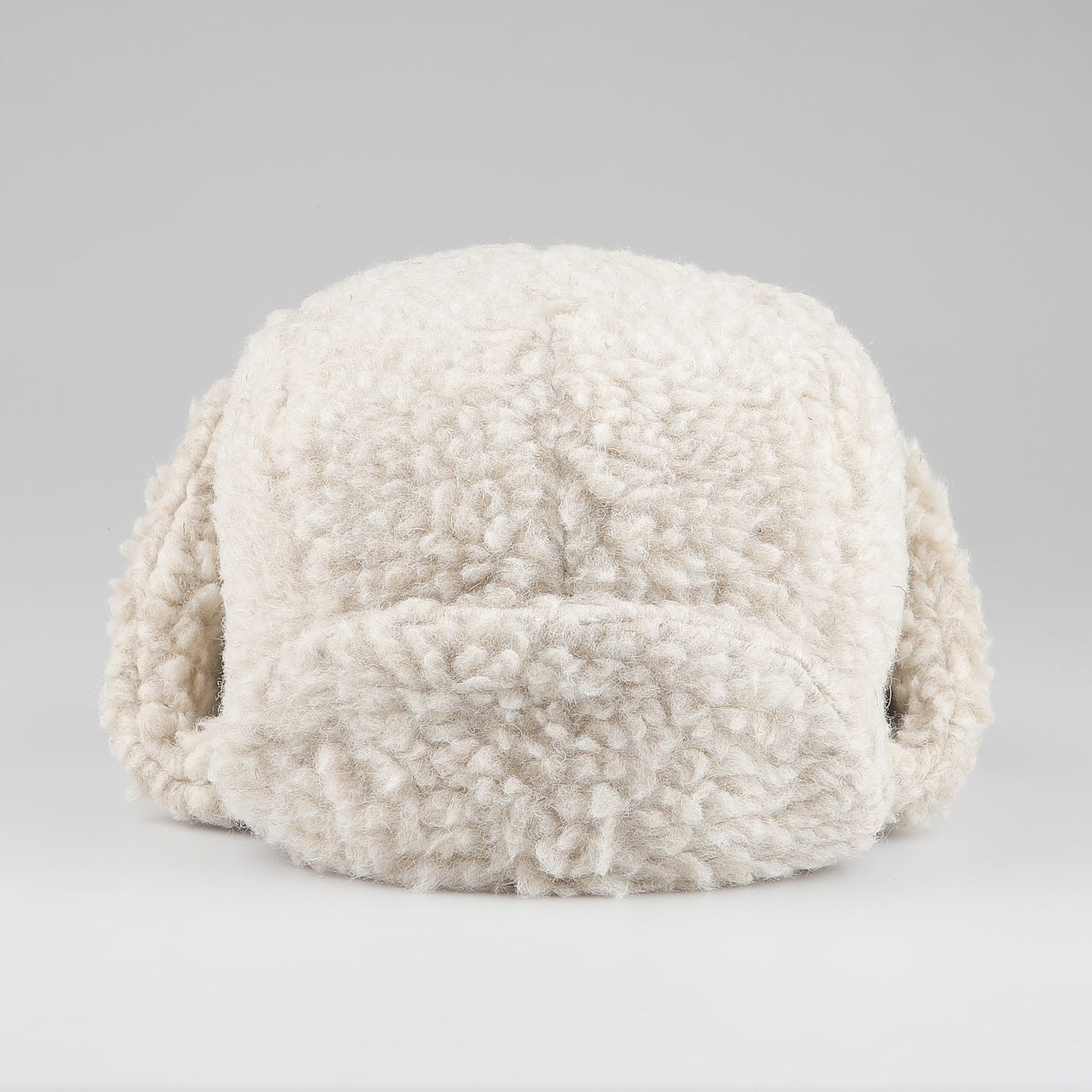 De Bonne Facture's Natural Wool Fleece Chapka Hat, featuring fluffy, cream-colored ear flaps, is shown from the front against a plain gray background.