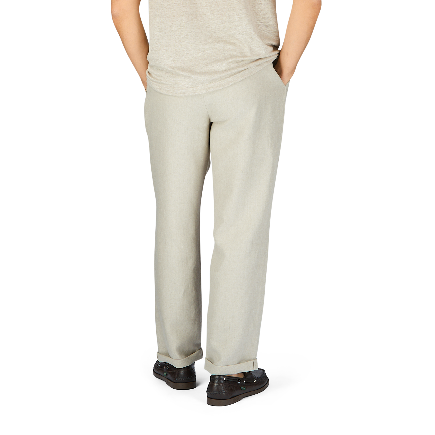 Rear view of a person in De Bonne Facture Oatmeal Beige Linen Pleated Trousers and gray shoes standing against a plain white background.
