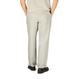 Rear view of a person in De Bonne Facture Oatmeal Beige Linen Pleated Trousers and gray shoes standing against a plain white background.
