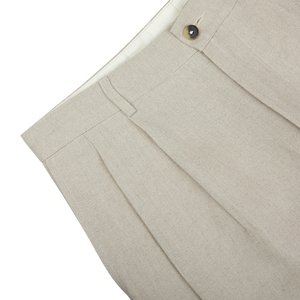 Close-up of De Bonne Facture's Oatmeal Beige Linen Pleated Trousers with a button and visible stitching against a white background.