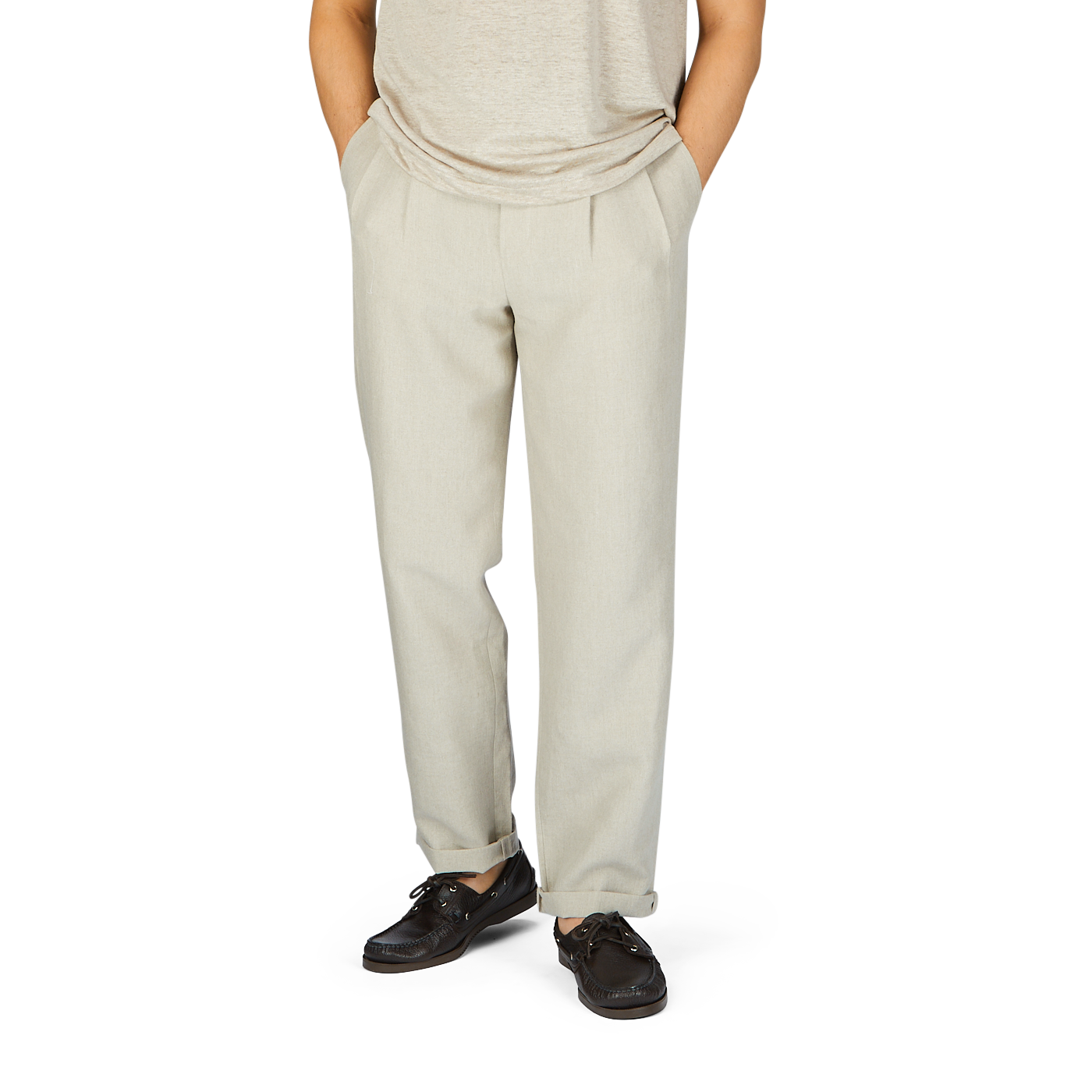 A person wearing De Bonne Facture's Oatmeal Beige Linen Pleated Trousers and brown leather shoes stands with hands in pockets against a plain background.