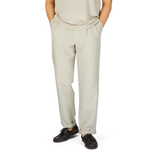 A person wearing De Bonne Facture's Oatmeal Beige Linen Pleated Trousers and brown leather shoes stands with hands in pockets against a plain background.