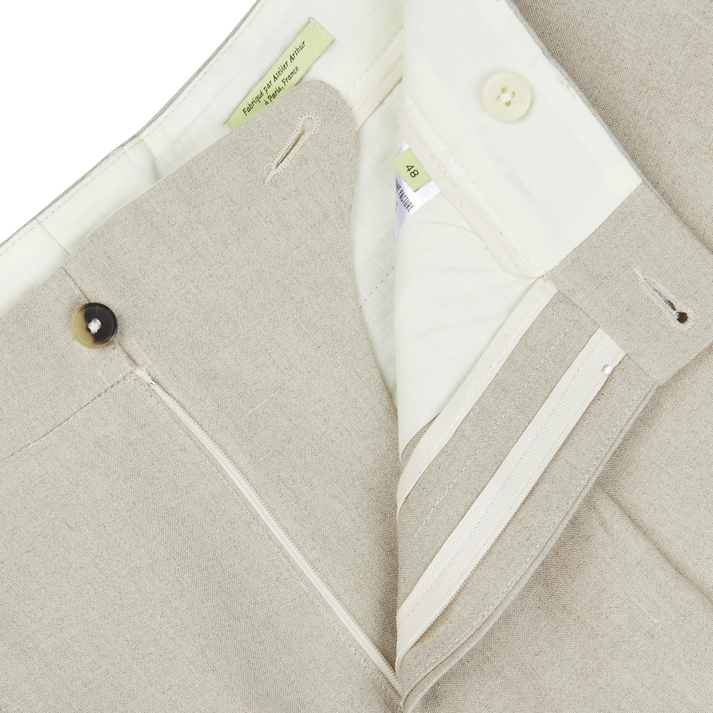 Close-up view of a contemporary fit oatmeal beige linen pleated trousers from De Bonne Facture with a button-up front, visible size tag, and collar details.