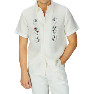 Man in a De Bonne Facture Off White Linen Floral Embroidered Shirt, standing against a grey background.