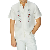 Man in a De Bonne Facture Off White Linen Floral Embroidered Shirt, standing against a grey background.
