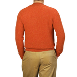 A person wearing a De Bonne Facture Orange Shetland Shaggy Dog Sweater and beige khaki pants, seen from behind. The background is plain and light-colored.