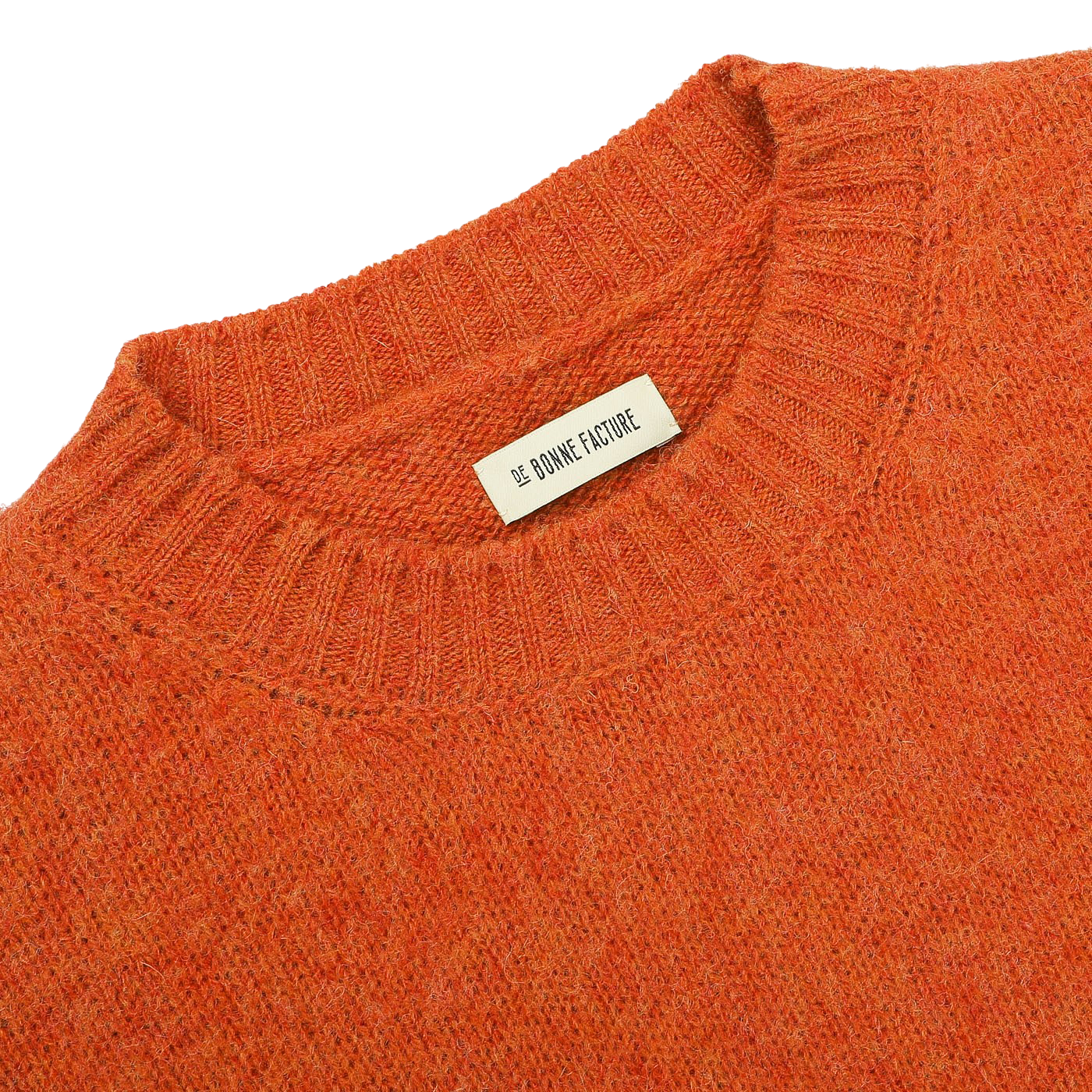 Close-up of an Orange Shetland Shaggy Dog Sweater with a ribbed neckline, featuring a tag that reads "De Bonne Facture." Made from pure Shetland wool, this regular-fit piece embodies warmth and style.