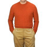 A person wearing the De Bonne Facture Orange Shetland Shaggy Dog Sweater and beige pants with hands in pockets stands against a plain background.