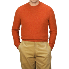 A person wearing the De Bonne Facture Orange Shetland Shaggy Dog Sweater and beige pants with hands in pockets stands against a plain background.