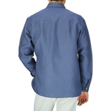 Back view of a man in a De Bonne Facture Pastel Blue Belgian Linen Two Pocket Overshirt and white pants against a grey background.