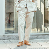 A person wearing De Bonne Facture's undyed heavy cotton drill balloon trousers and a relaxed fit, light beige suit with tan shoes standing in front of a glass building, cropped at mid-torso.