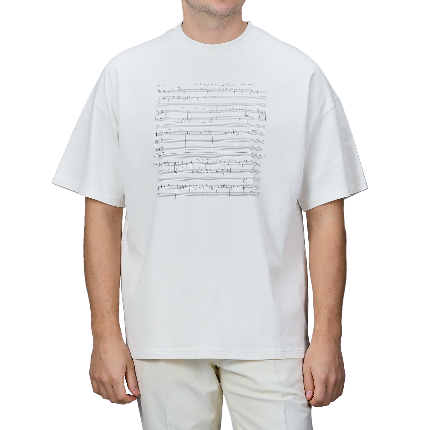 A person is wearing the Undyed Printed Cotton Jersey T-Shirt by De Bonne Facture, which features a music sheet design on the front and is made from organic cotton jersey. The background of the T-shirt is plain gray.