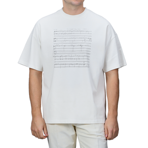 A person is wearing the Undyed Printed Cotton Jersey T-Shirt by De Bonne Facture, which features a music sheet design on the front and is made from organic cotton jersey. The background of the T-shirt is plain gray.