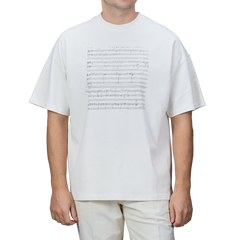 A person is wearing the Undyed Printed Cotton Jersey T-Shirt by De Bonne Facture, which features a music sheet design on the front and is made from organic cotton jersey. The background of the T-shirt is plain gray.