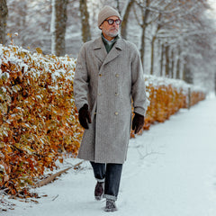 A man in a De Bonne Facture Undyed Wool Herringbone Grandad Coat walking in the snow.