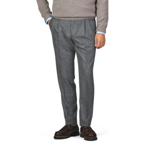 A person dressed in a gray sweater, De Petrillo's Medium Grey Wool Flannel Modello B Trousers, and dark brown shoes stands with their hands in their pockets.