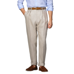 A person wearing De Petrillo Sand Beige Irish Linen Modello E Trousers, a brown belt, a light blue shirt, and brown loafers stands confidently with hands in pockets, exuding casual elegance.