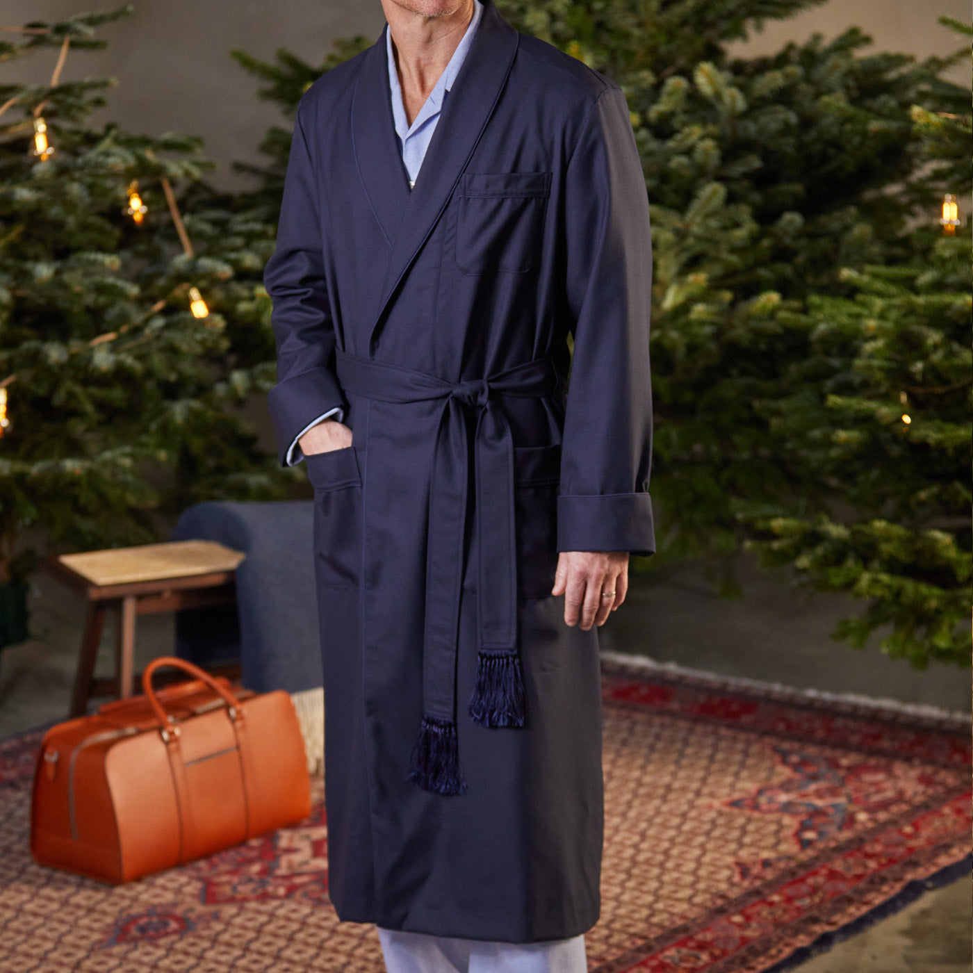 A man is standing in front of a Christmas tree wearing a Derek Rose Navy Blue Wool Twill Westminister Gown.