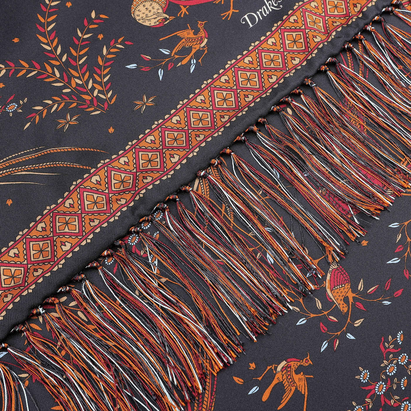 A close-up of the Navy Birds of Paradise Print Tubular Silk Scarf by Drake's showcases a fringed edge with a colorful and intricate pattern featuring animals, leaves, and geometric designs on a dark background. This exquisite scarf draws inspiration from the rich textile traditions of Como, Italy.