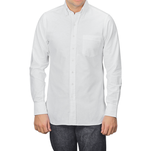 A man in a Drake's White Classic Cotton Oxford BD Shirt with a pocket on the left and dark jeans stands against a gray background.