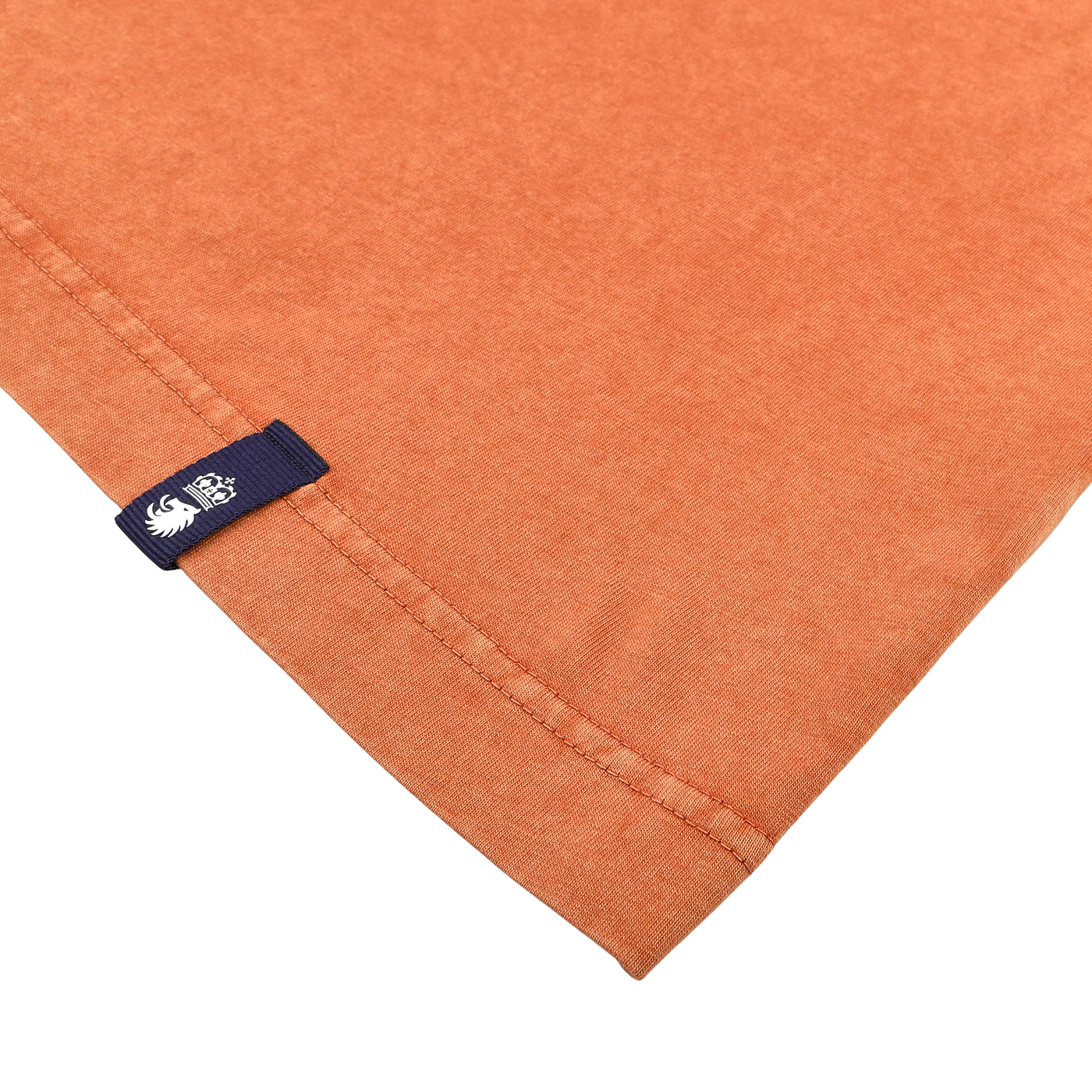 Close-up of the hem of a Drumohr Burnt Orange Washed Cotton T-Shirt, highlighting a stitched edge and small blue fabric tag with a design, laid on a white surface.