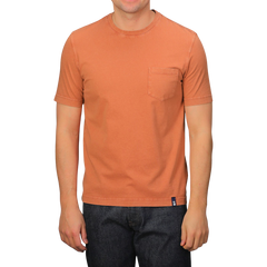 A person dons a Drumohr Burnt Orange Washed Cotton T-Shirt, featuring a slim fit and chest pocket, crafted in Italy, paired with dark jeans.