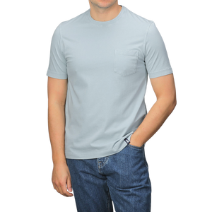 A person in a slim-fit, light gray Drumohr T-shirt with a chest pocket and sky blue jeans stands with one hand in their pocket.