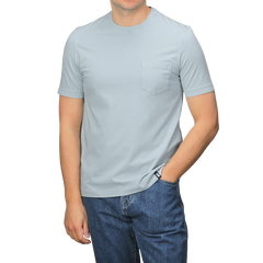 A person in a slim-fit, light gray Drumohr T-shirt with a chest pocket and sky blue jeans stands with one hand in their pocket.